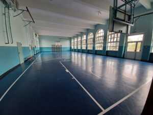 palestra della scuola primaria di villafranca. Le pareti sono di colore bianco e azzurro, il pavimento blu con le strisce del campo bianche. Sul soffitto sono appesi dei canestri.